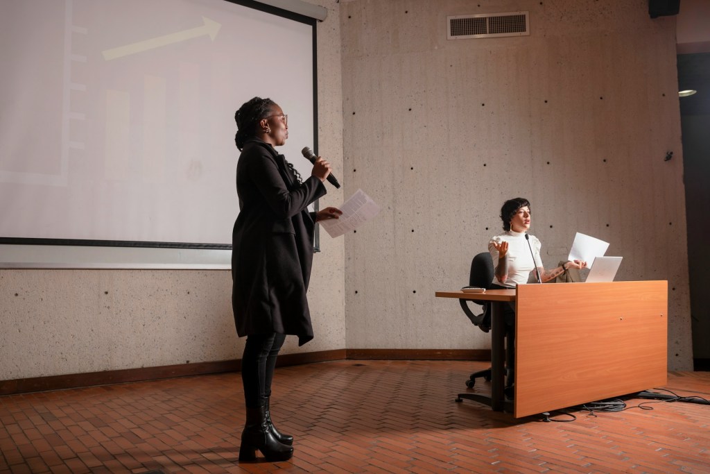 A black woman at the front of a large lecture hall holds a microphone in discussion with a white  woman with short brown hair sitting at a desk nearby, also at the front of the room. That woman is speaking near a smaller lectern microphone. Each woman holds papers in her hands.