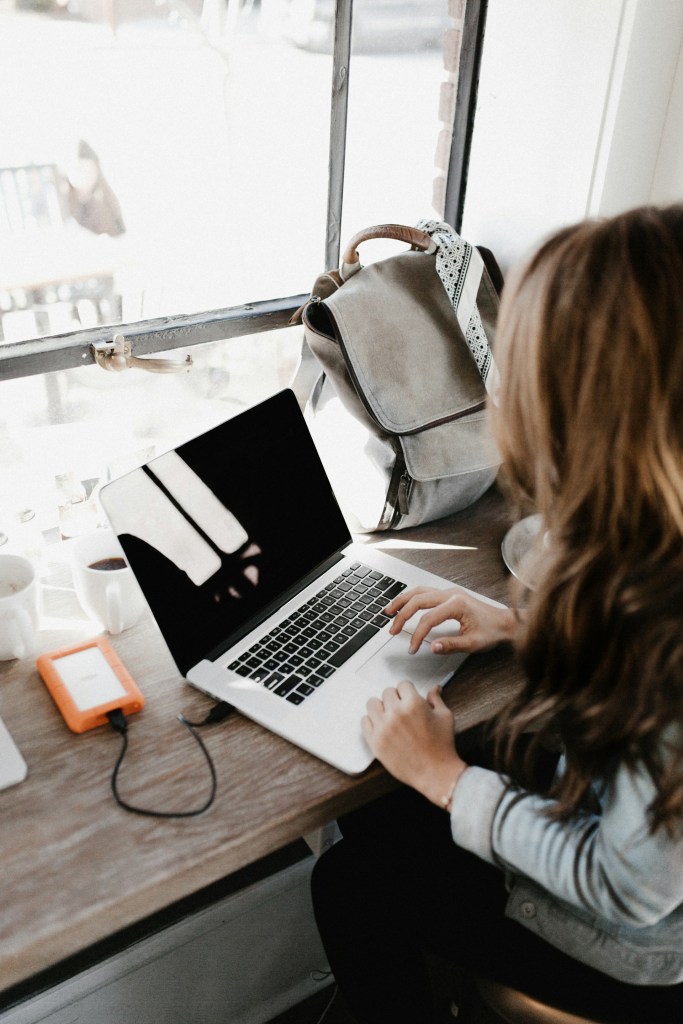 A woman looking at an open laptop on a desk in a coffee shop. Her backpack is leaning in the corner by the window. It's a sunny day, with light streaming in.