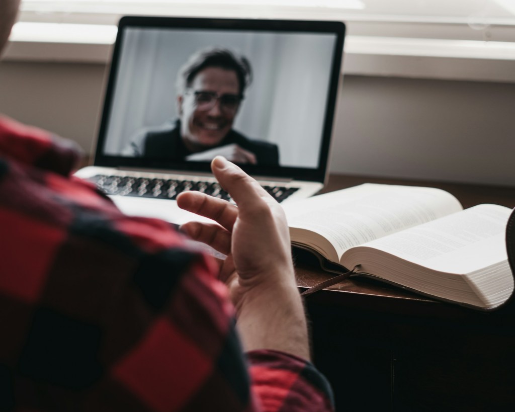 View over the shoulder of a professor who is attending a live virtual event. On his open laptop screen is the speaker, a person with short hair and glasses, smiling.