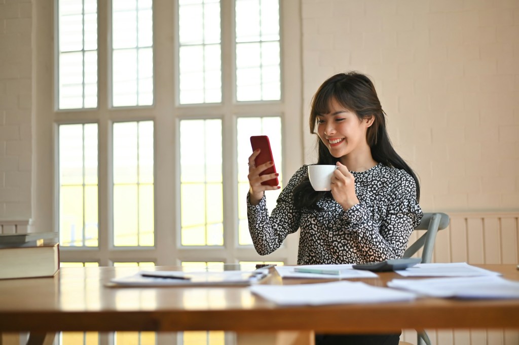 A bright room where a woman sits at a table with papers. She's holding an small mug in her hands and is smiling at the red mobile phone in her hands.