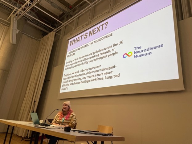 Image 6: Estonian-UK PhD candidate Iris Sirendi at a long desk speaking to a projection with a slide with the heading ‘What’s Next?’ and a logo that reads ‘The Neurodiverse Museum’.