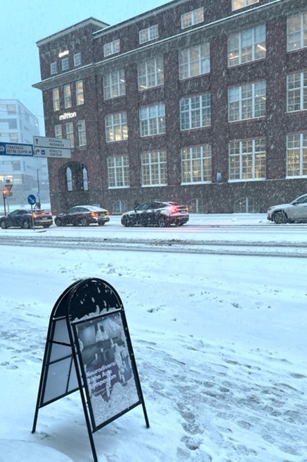 Image 9: A signboard ‘Neurodiversity in the Arts Symposium’ covered in snow, in a street raging with a snow-storm with cars passing by in front of a building across the road.