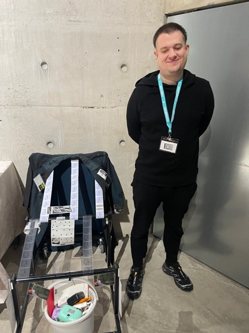 Image 8: Dr Fran Trento standing next to their mobile installation that comprises a jacket onto which participants can make marks onto, scrolls of film, and a pail with cameras and other creative and critical tools to dismantle harmful narratives and approaches