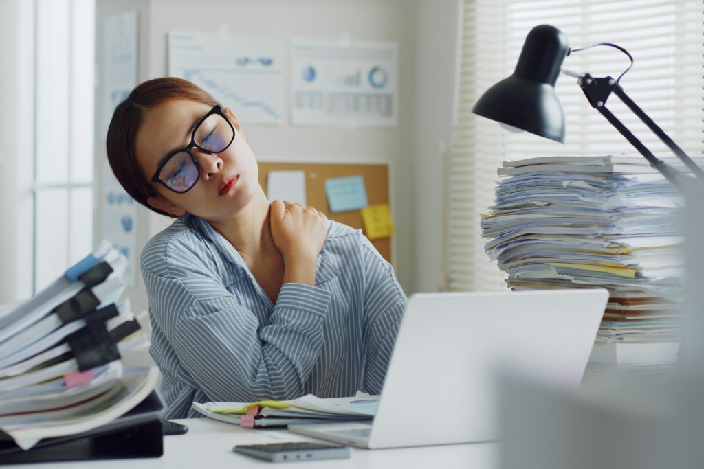 An Asian woman stretches her neck while sitting at a desk surrounded by stacks of papers. She seems tired and burntout.