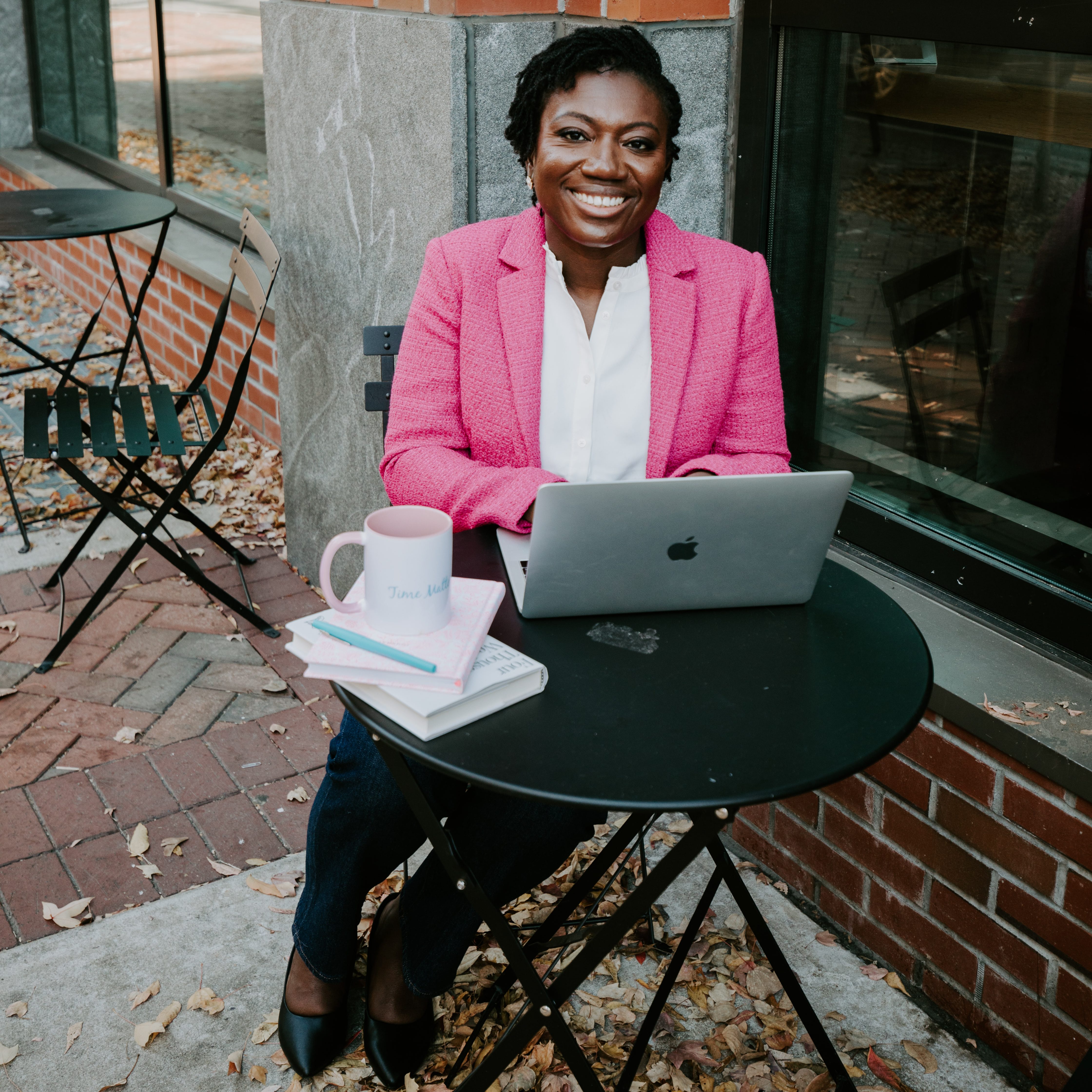 Dr. Martha Kenney in a bright pink blazer, white blouse, and dark pants sits at a round table outside in the fall by a brick building and walkway