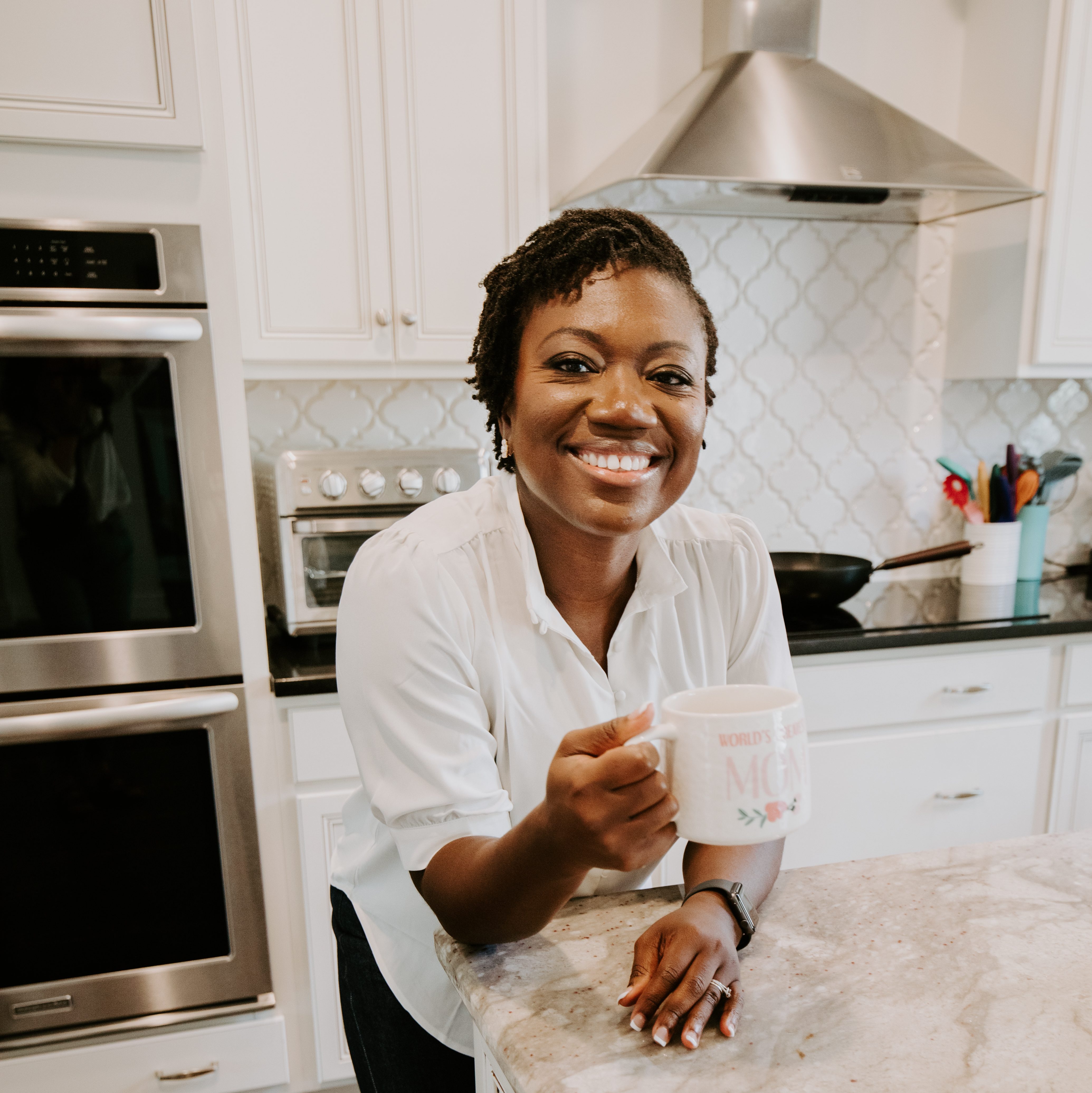 Dr. Martha Kenney holds a mug that says 'world's best mom' while leaning on her kitchen counter. Behind her is a double oven, an air fryer, and an induction stovetop with a hood.