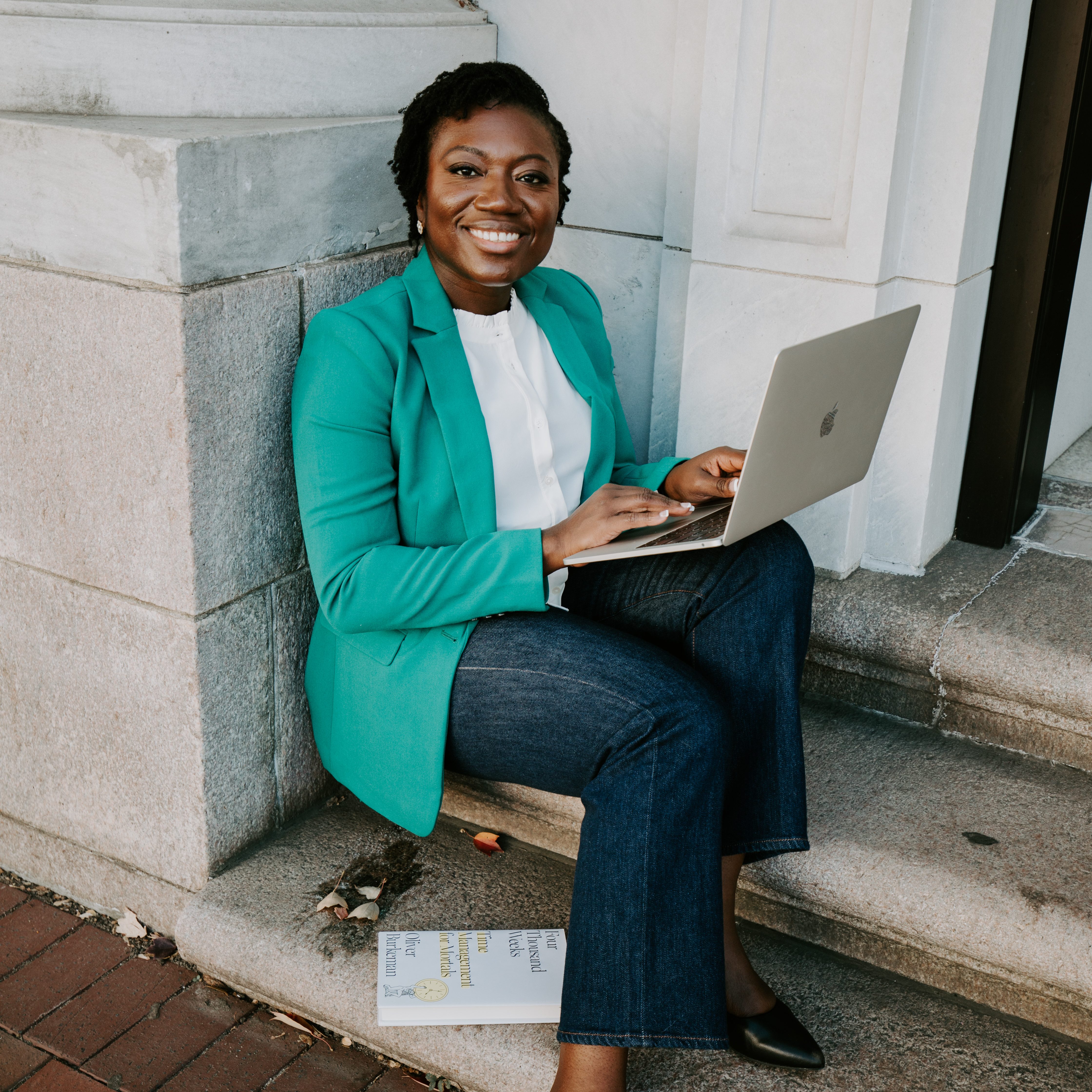 Dr. Martha Kenney in a turquoise blazer and dark jeans sits on low stone stairs outside a building entrance with an open Apple laptop on her lap. She's smiling.