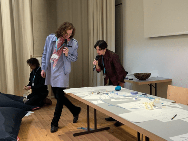 Image 5a: shy*play’s Antje Nestel and Aion Arribas, both holding microphones and reading from papers strewn on a long table