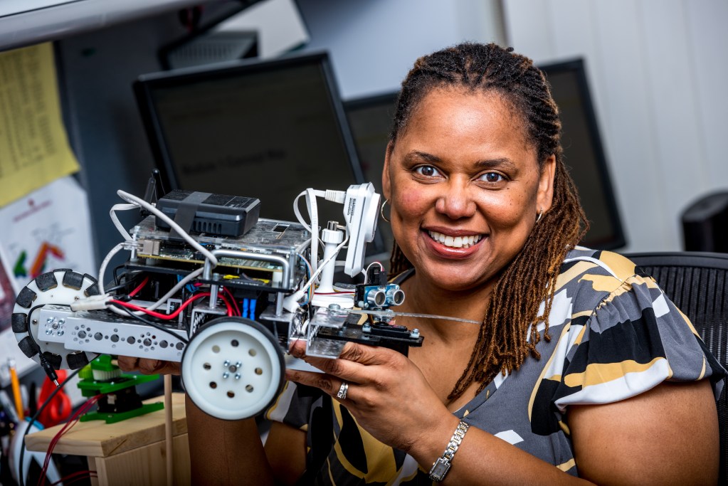 Dr. Carlotta Berry, a professor of robotics and engineering, holds a robot with 3 wheels in her hands. She's smiling at the camera in her university office.