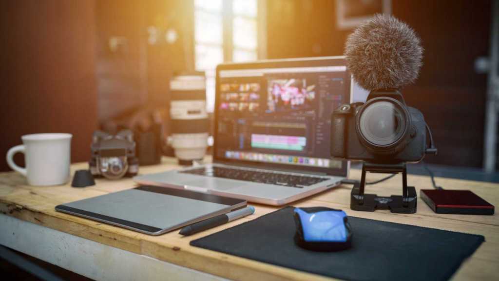 A video recording set-up with a professional camera and microphone, an open laptop with a video editor on the screen, and a mug.