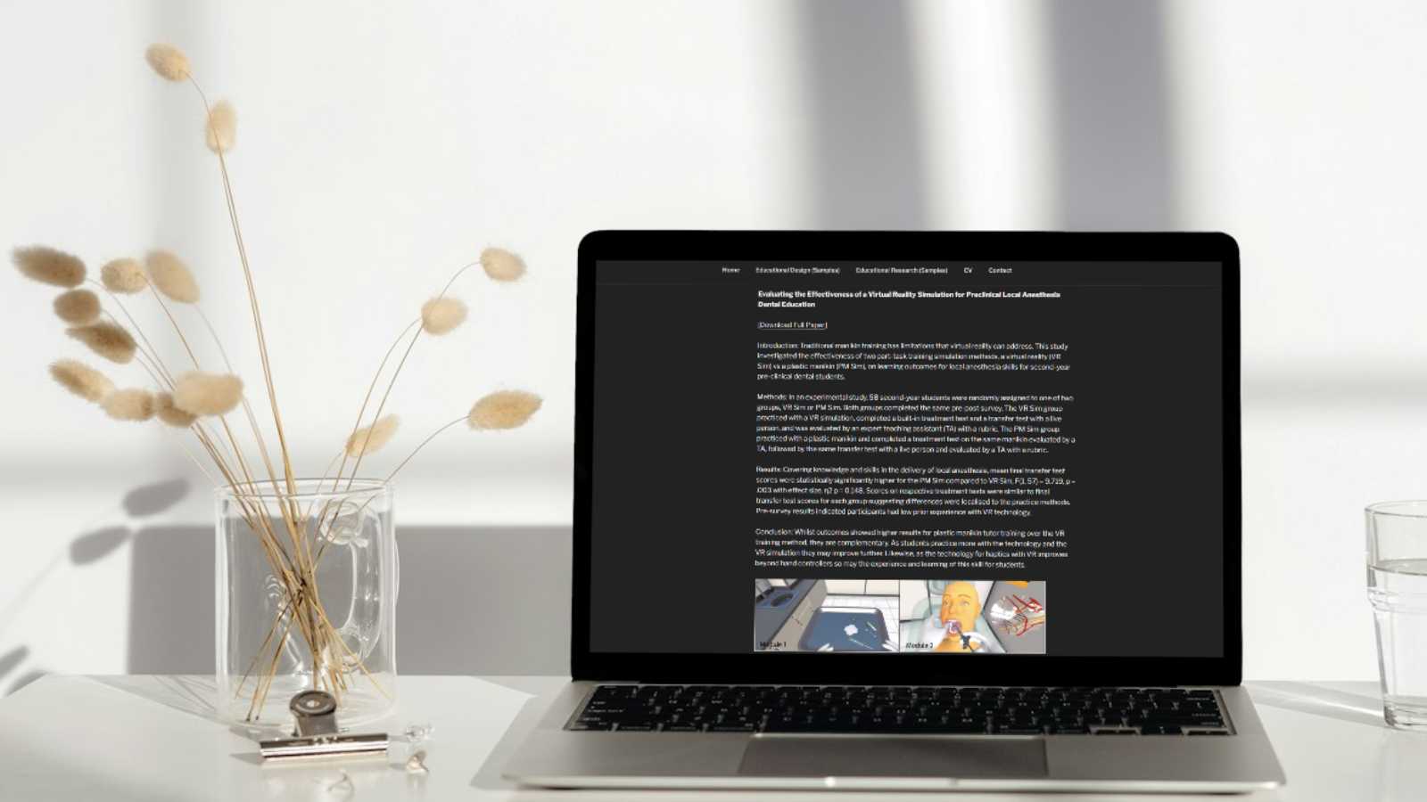 An open laptop on a white desk next to a clear glass mug holding sprigs of dried decorative plants with puffed ends. There is a clip holder and a glass of water also on the table. On the open laptop screen is Dr. Elizabeth McAplin's website with her Educational Research page pulled up.