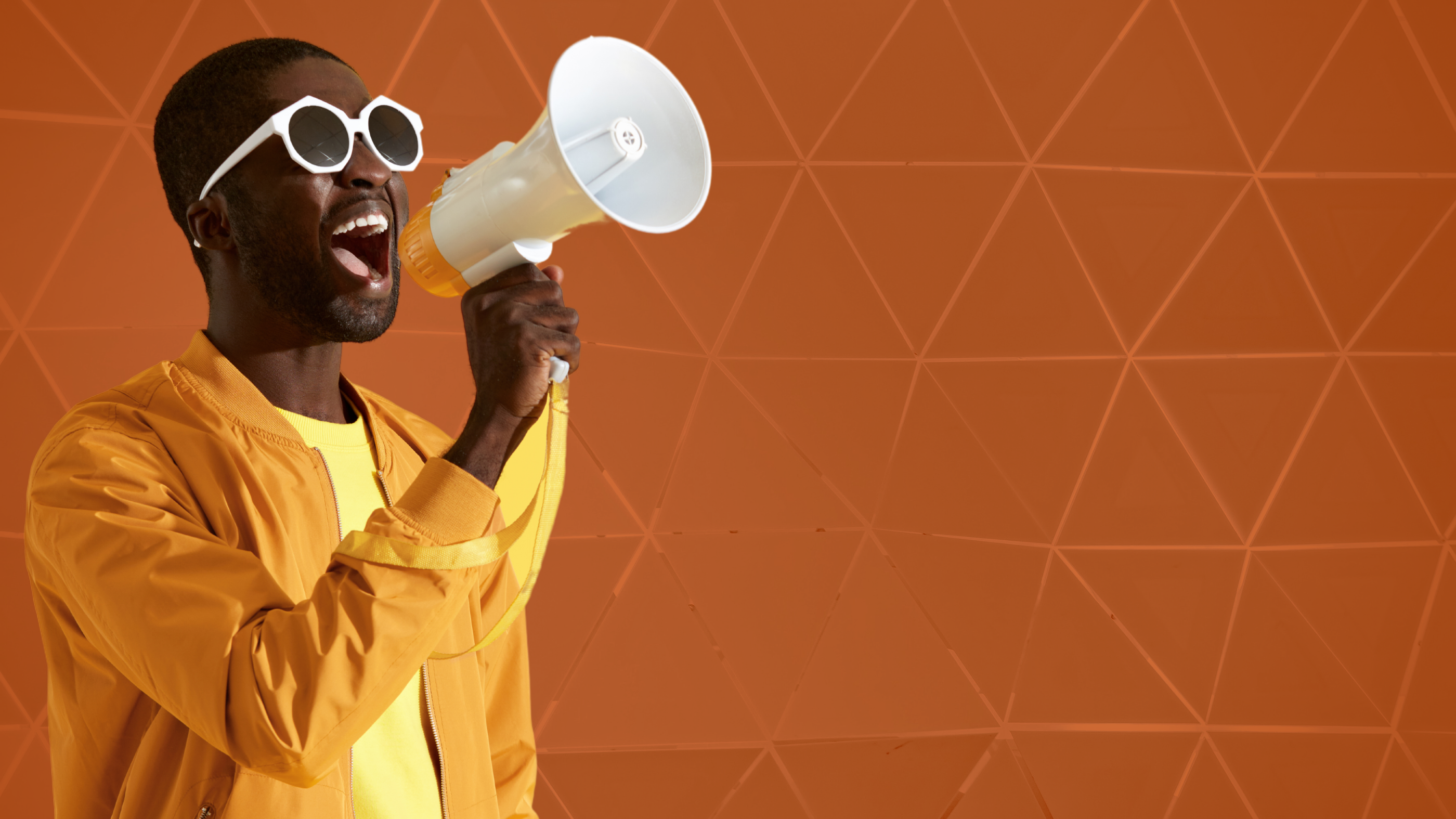 A black man in a yellow outfit holds a speaker horn. His mouth is open to speak an announcement.