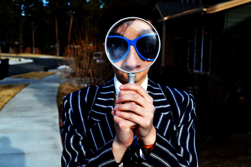 An Asian man in a pinstripe suit holds a large magnifying glass in front of his face. He is wearing blue sunglasses that look funny because they are so large in the magnifying glass to the viewer.