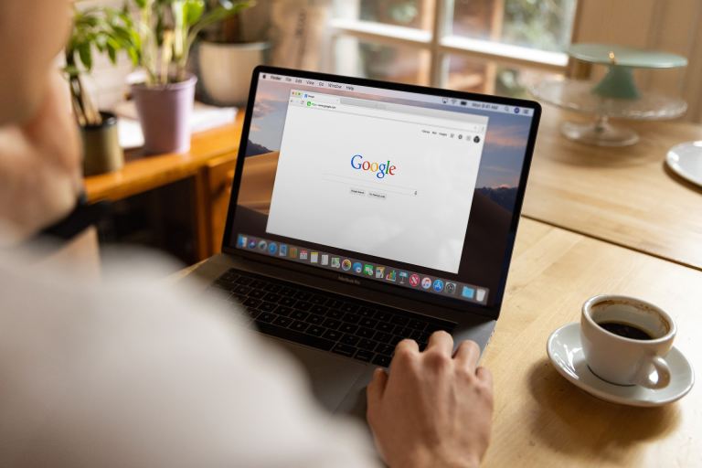 A white person sits at a table with their laptop and a cup of coffee on a saucer. The open laptop has an internet browser with Google Search pulled up.