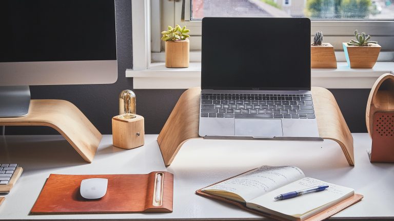 A desk workspace with a propped up monitor and laptop screens. On the desk are a mouse and an open notebook with a pen.