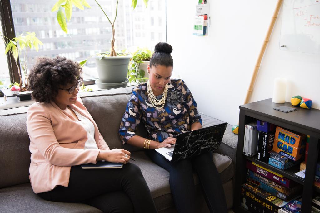 Two black women sit on a gray sofa in on office. They are looking at a laptop and smiling.