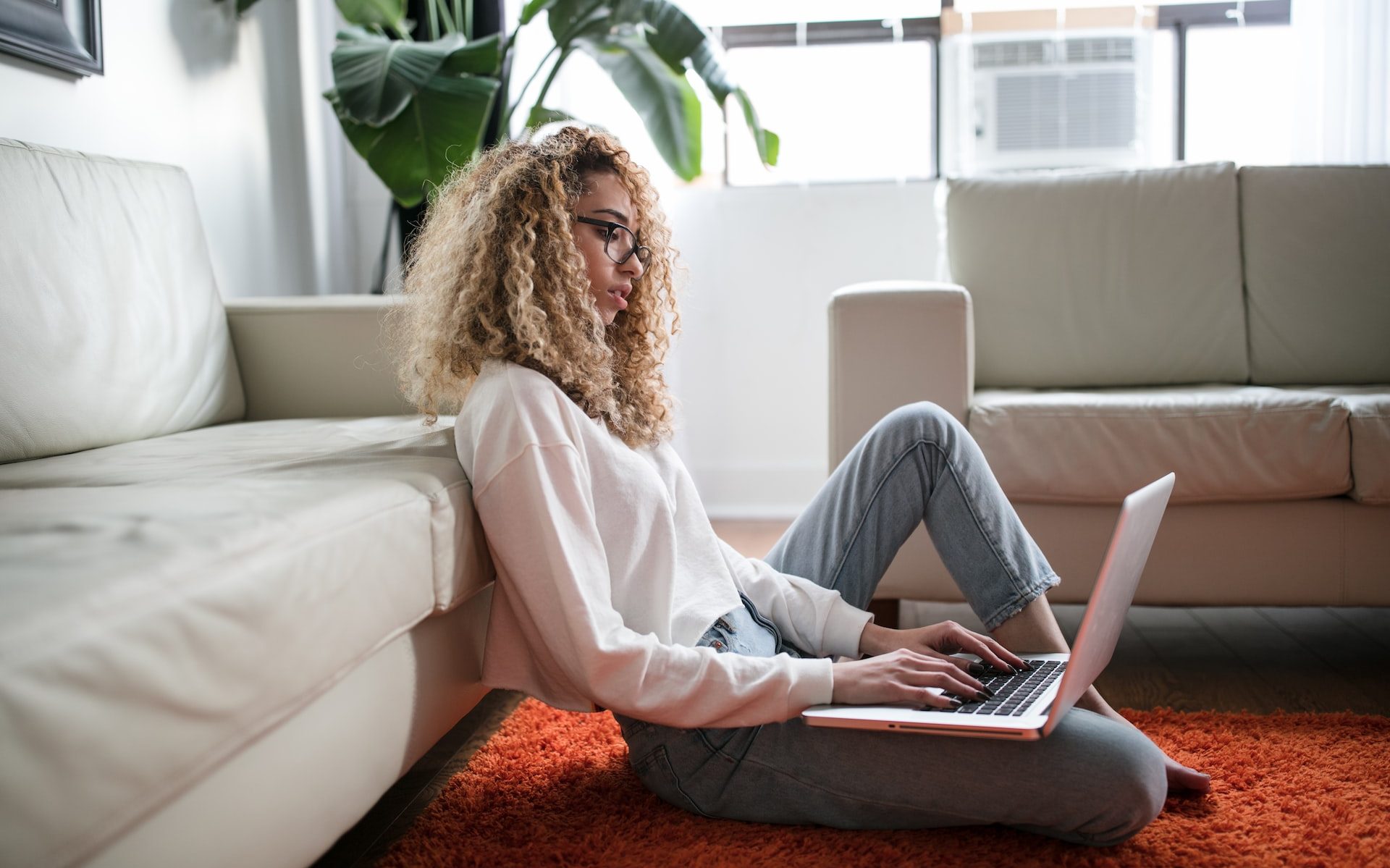 A black woman sits on a plush orange rug leaning against a tan sofa. She is typing on her laptop which rests on her knee which is bent under her.