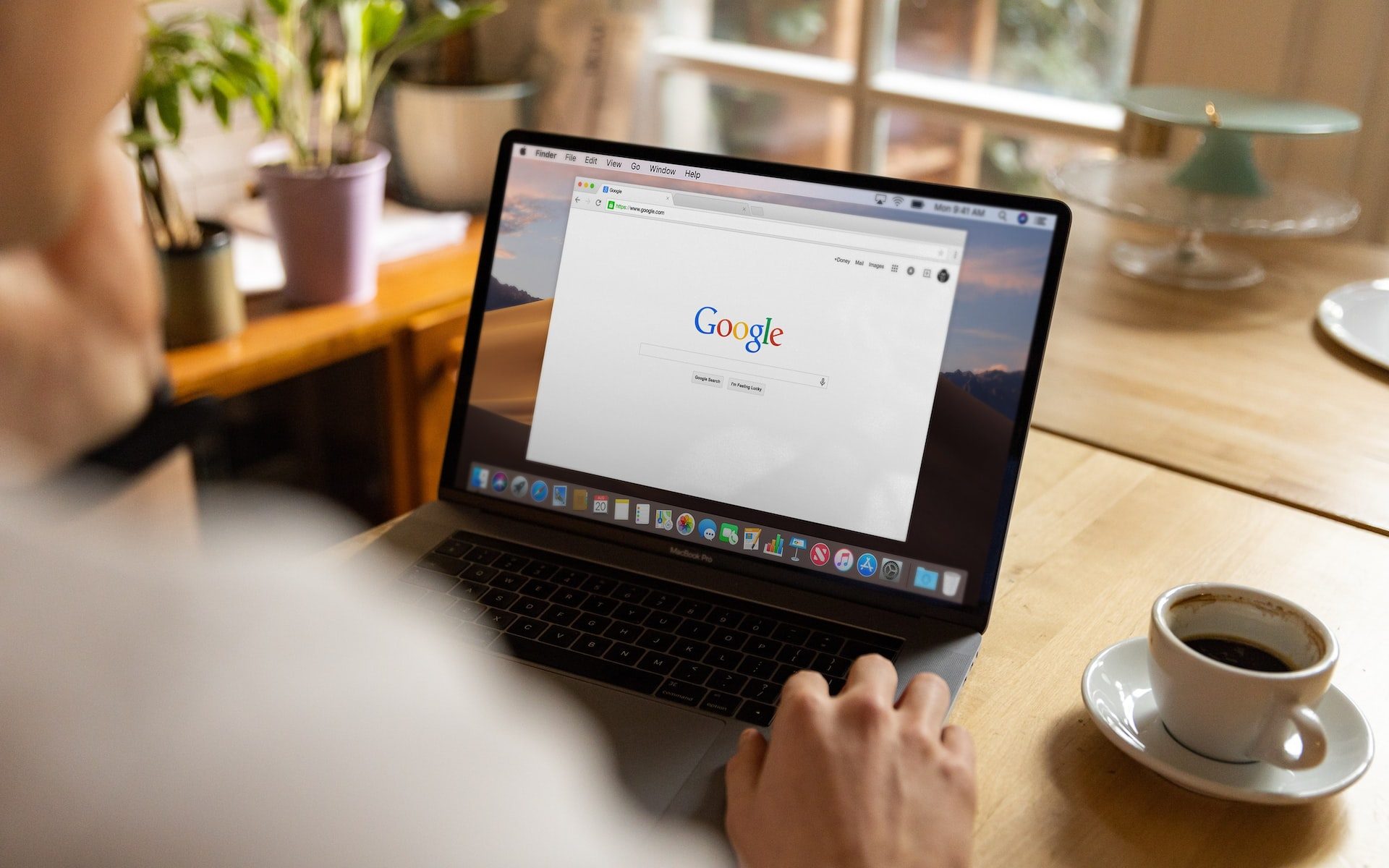 A person sits at their kitchen table with an open laptop and a cup of coffee. On the laptop screen is a browser with the Google Search page pulled up.