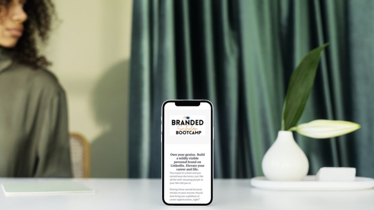 A mobile phone standing on a desk. On the phone's screen is info about Dr. Gertrude Nonterah's course, The Branded Scholar Bootcamp. In the background of the photo, a young black woman is sitting at the desk. On the desk is a small white vase with a leafy plant in it. There are long green curtains covering a window behind the desk.