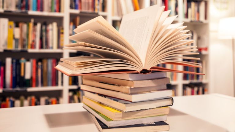 A stack of books on a table. The top book on the stack is open. Behind the stack of books are library shelves.