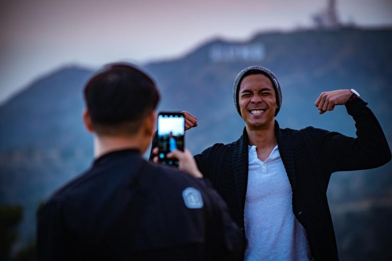 A man holds a phone taking a photo of his friend who is smiling. They are in Los Angeles. The Hollywood sign is visible in the background, but unreadable because the background of the photo is very blurry.