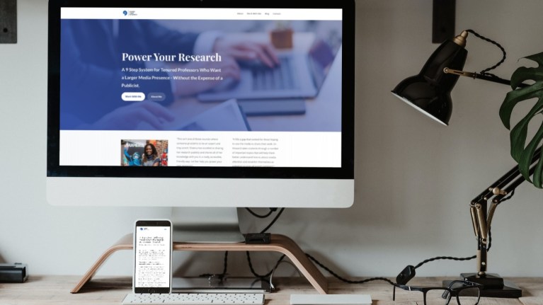 A desktop screen with the Power Your Research program pulled up. Propped up against the monitor riser is a mobile phone with one of Dr. Howard's blog posts. On the desk is a keyboard, glasses, and a black and gold desk lamp turned off.