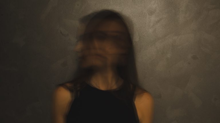 A photo of a woman blurred, probably by having the camera take a long still photo while the subject, a woman with brown hair, moves her head back and forth. This photo represents the internal decision women  have to make when being asked to do free speaking engagements.