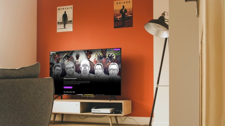 A television with the Milestone Generations (2022) documentary from HBO Max is pulled up on the screen. The tv is on a small blondewood modern tv stand. Behind the tv is an orange accent wall with two posters. In front of the TV is a gray couch. In the corner of the room is a tall black floor lamp, turned off.