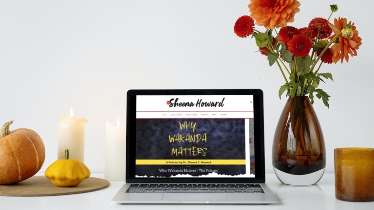 The personal website of Dr. Sheena Howard on a laptop screen next to a fall flower arrangement in a vase, and some fall decorations like a candles, and two small pumpkins.