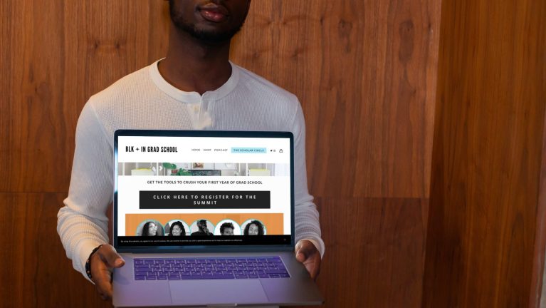 A black man wearing a long sleeved white Henley shirt holds an open laptop  against his chest presenting the screen toward the camera. On the laptop screen is information about the Grad School Success Summit from the Blk + In Grad School website.