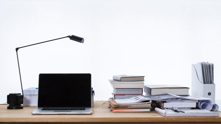 A desk with an open laptop, small reading light, piles of books and papers.