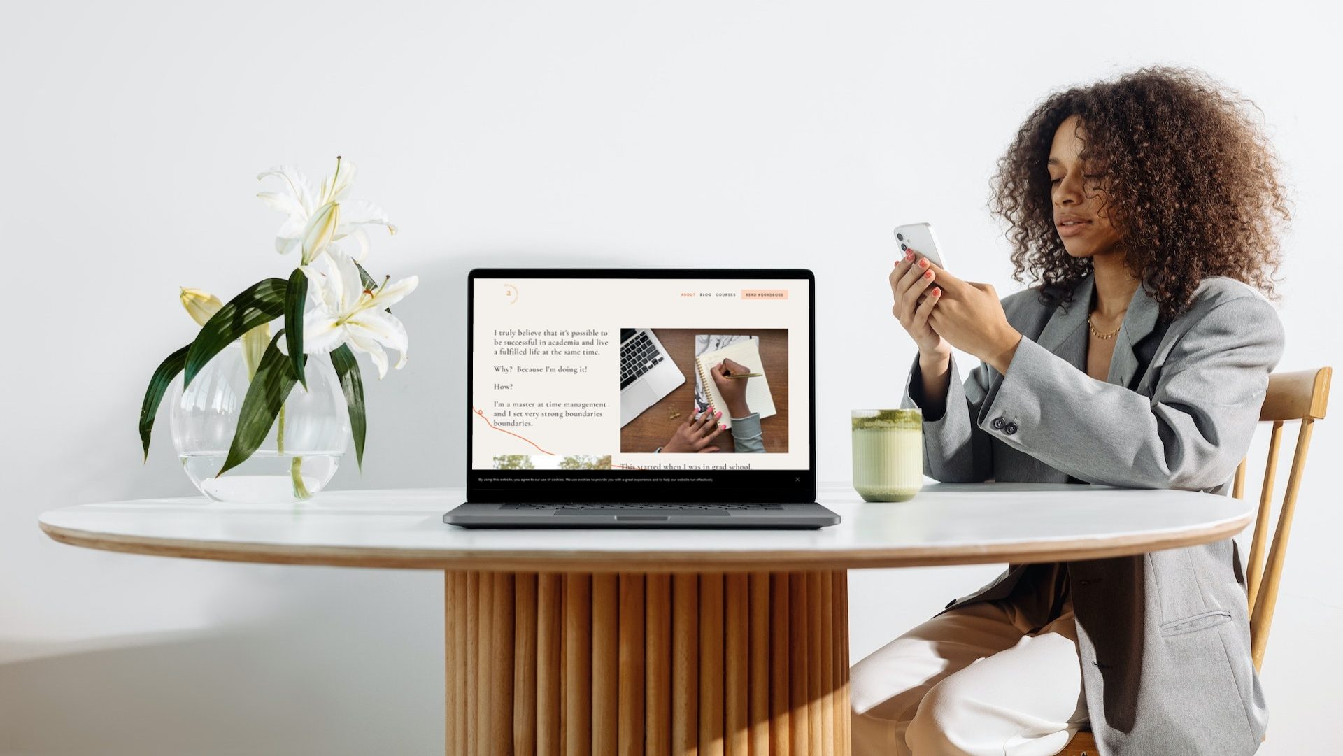 A black woman wearing an oversized gray blazer and white slacks sits at a round modern table with a cup of coffee, holding her mobile phone. Also on the table are a vase of lilies, and an open laptop with The Academic Society website pulled up.