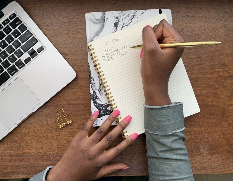 Toyin Alli holding a hold pen writing a to-do list in a spiral bound notebook. On the list are email, announcements, solutions, and office hours. Next to the notebook is a gold binder clip and a laptop, mostly out of shot.