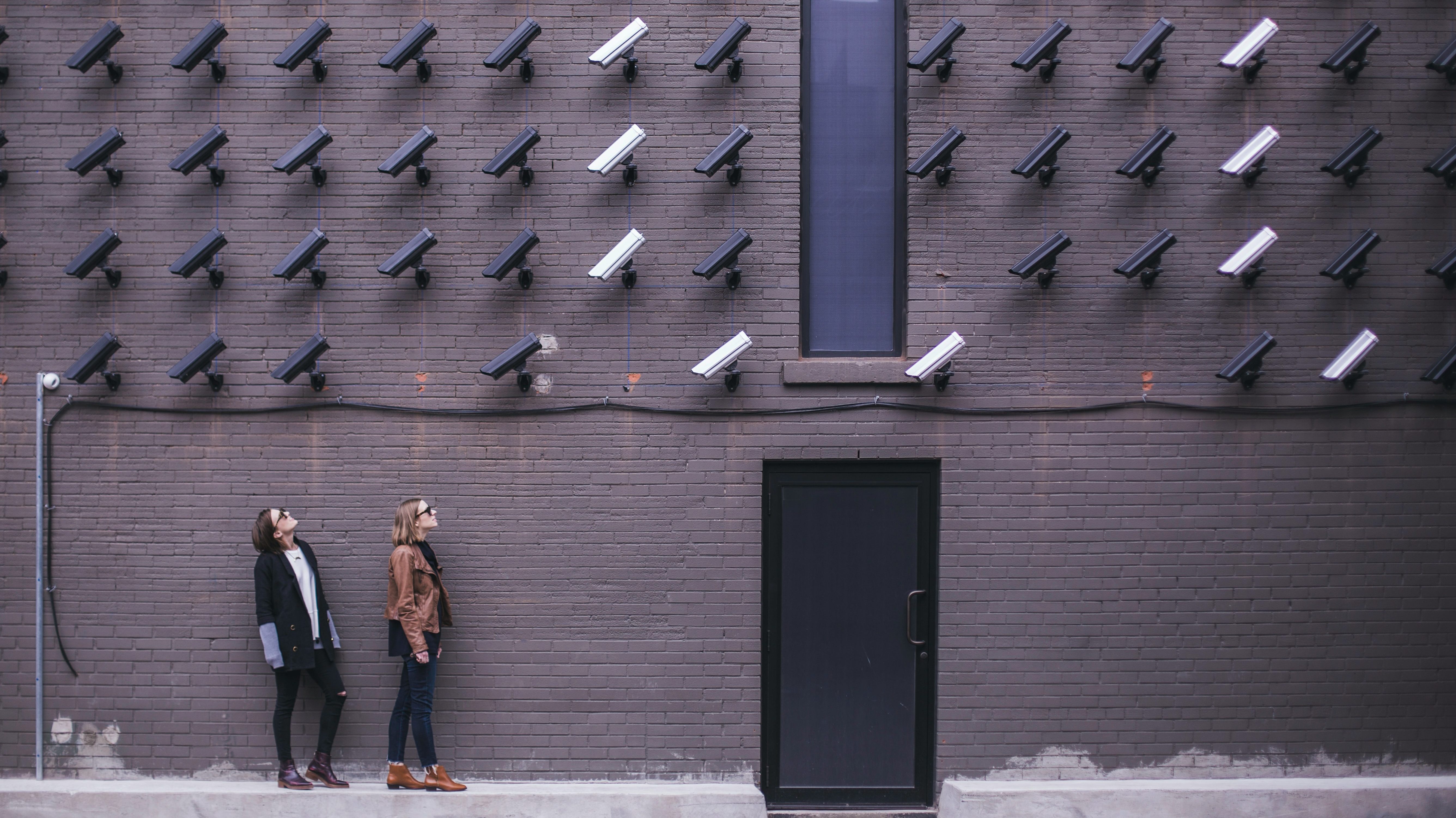 Two women stand on a raised sidewalk wearing booties, pants, jackets, and sunglasses. They are both looking up towards dozens of video surveillance cameras on the brick wall above them. The cameras all point down making it feel hyper-surveilled.