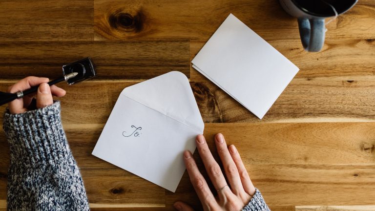 Woman wearing blue heathered sweater dips a fountain pen into ink. She is writing the address on an envelope to send in the mail.