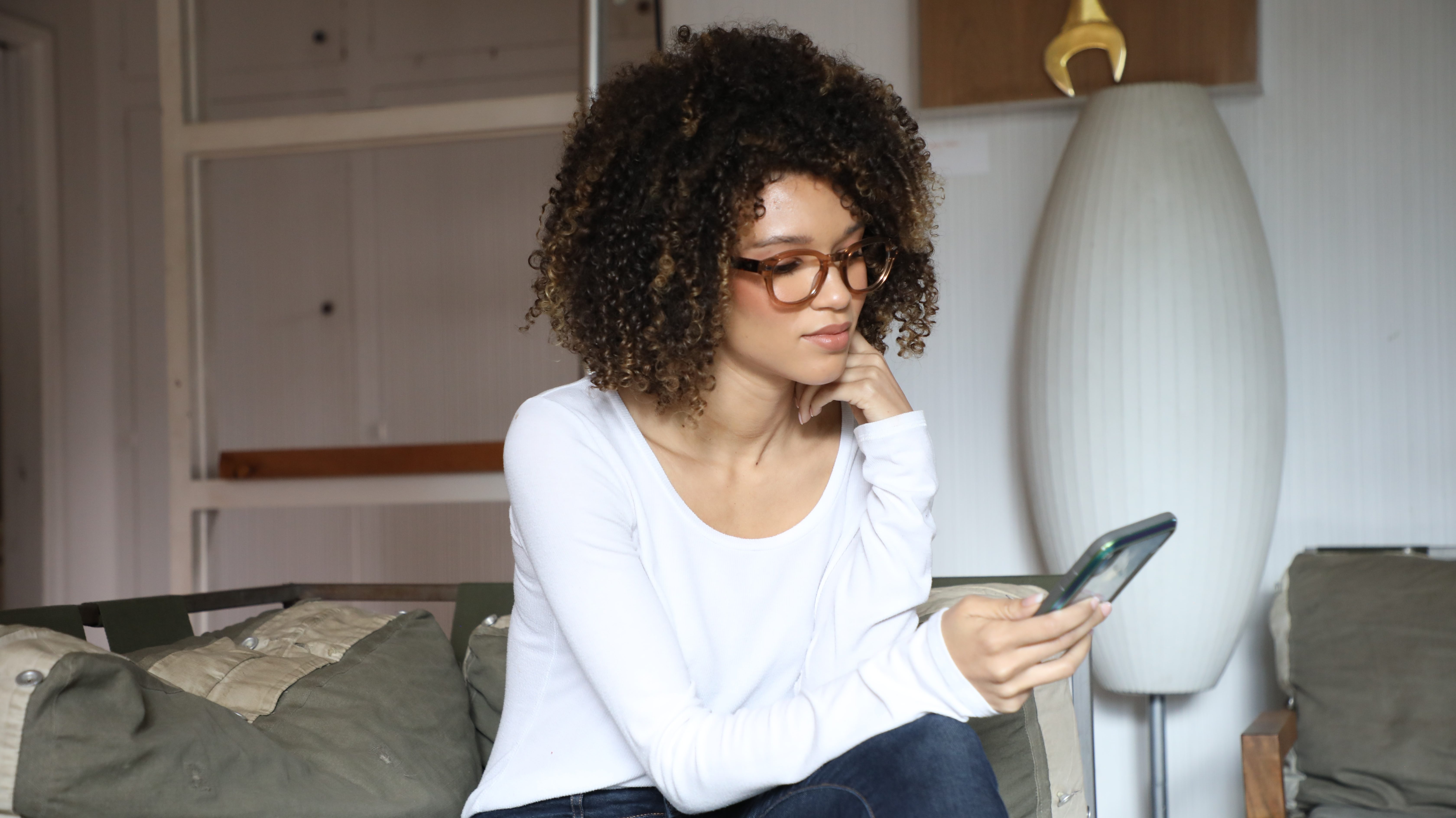 Black woman with brown square glasses and a long sleeve white t-shirt looks at her phone whiles sitting on a couch.