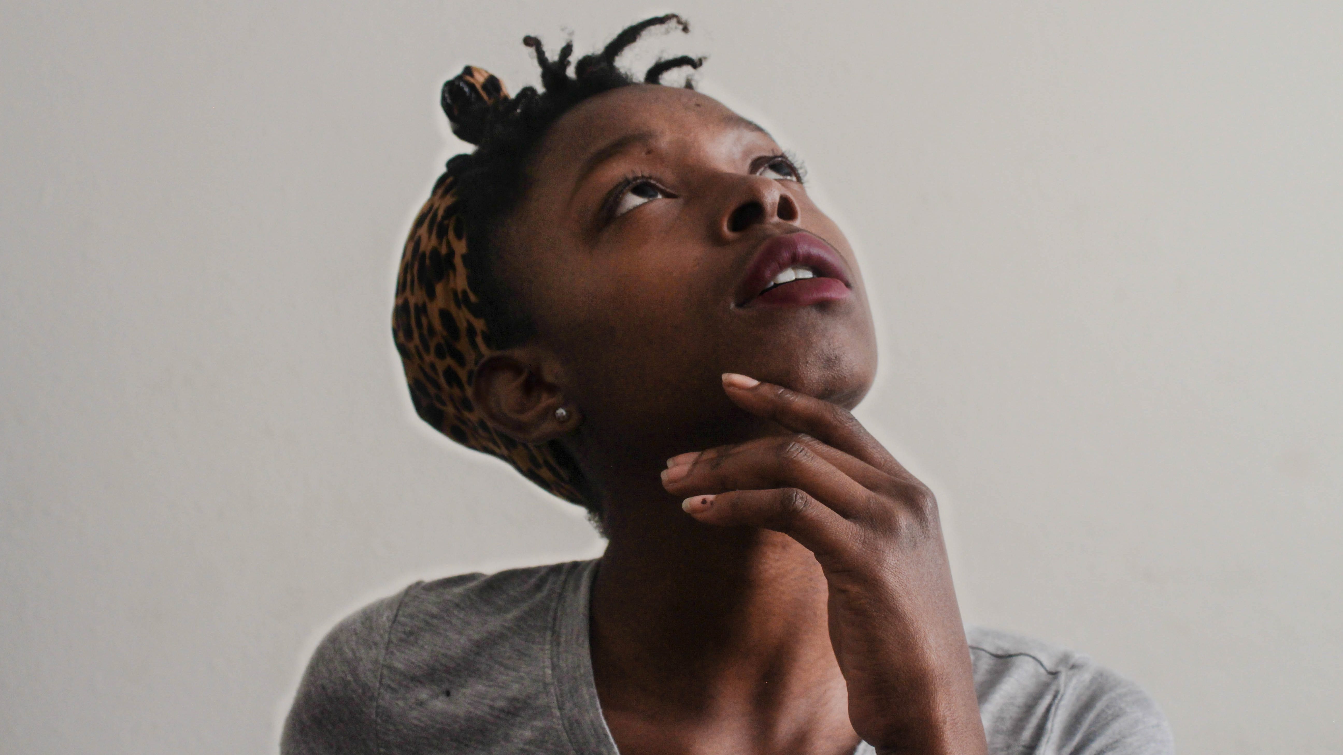 Black woman wearing gray t-shirt and cheetah headwrap holds her hand to her chin and looks up, as if pondering.