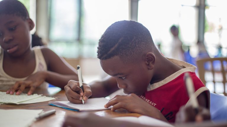 A black elementary school boy and girl sit at a table working. The boy is wearing a red sleeveless Miami Heat shirt and holding a pencil. They are both concentrating. Photo by Santi Vedri.