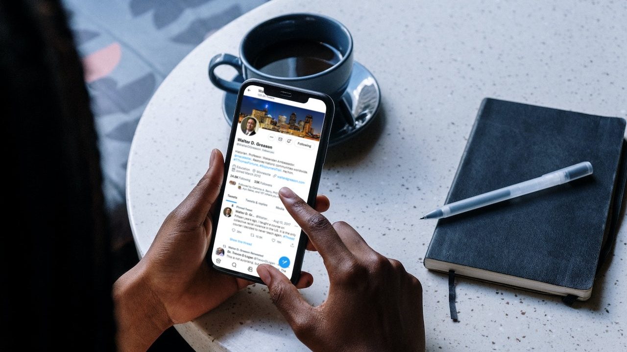 A black man holds an iPhone 11 at a small round table with a coffee cup and journal on top of it. On the phone screen is the Twitter profile of @WalterDGreason.