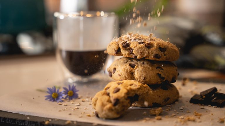 Three chocolate chip cookies are stacked on top of each other next to a cup of espresso in a clear glass. There are two small purple flowers next to the cookie.