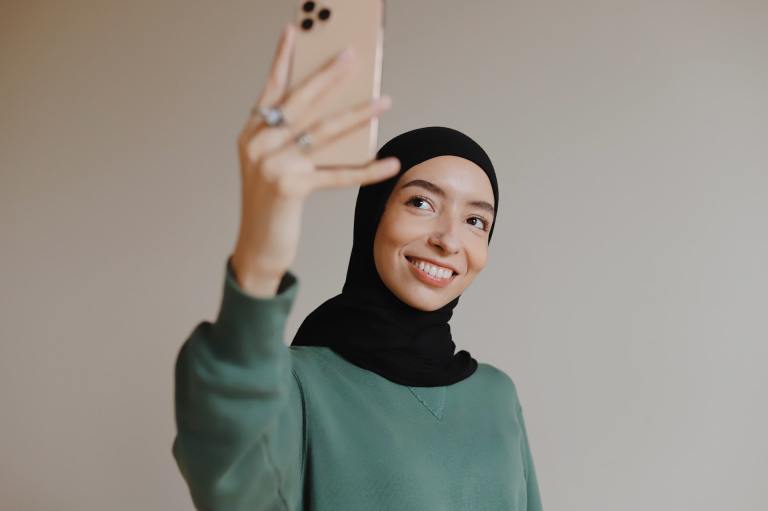 A woman wearing a hijab and a green sweater holds her phone and smiles at the camera while taking a selfie photo.