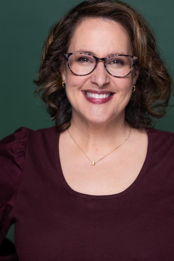 Jasmine Bucher smiles for a headshot wearing brown-framed glasses and a dark blouse. She is standing in front of a dark green backdrop and has curly brown shoulder-length hair