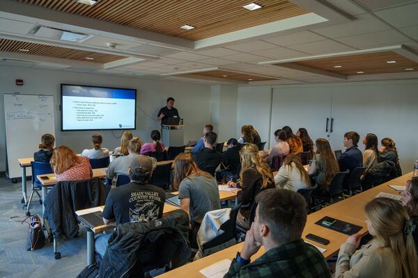 Students face the front of a classroom while a speaker presents using an electronic screen.