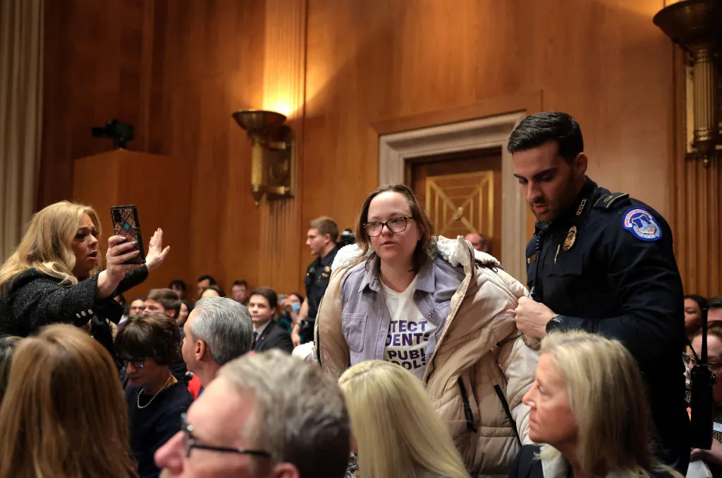 A person is standing near another person in a uniform. The room is full with other people sitting. Another person is standing and holding a cell phone