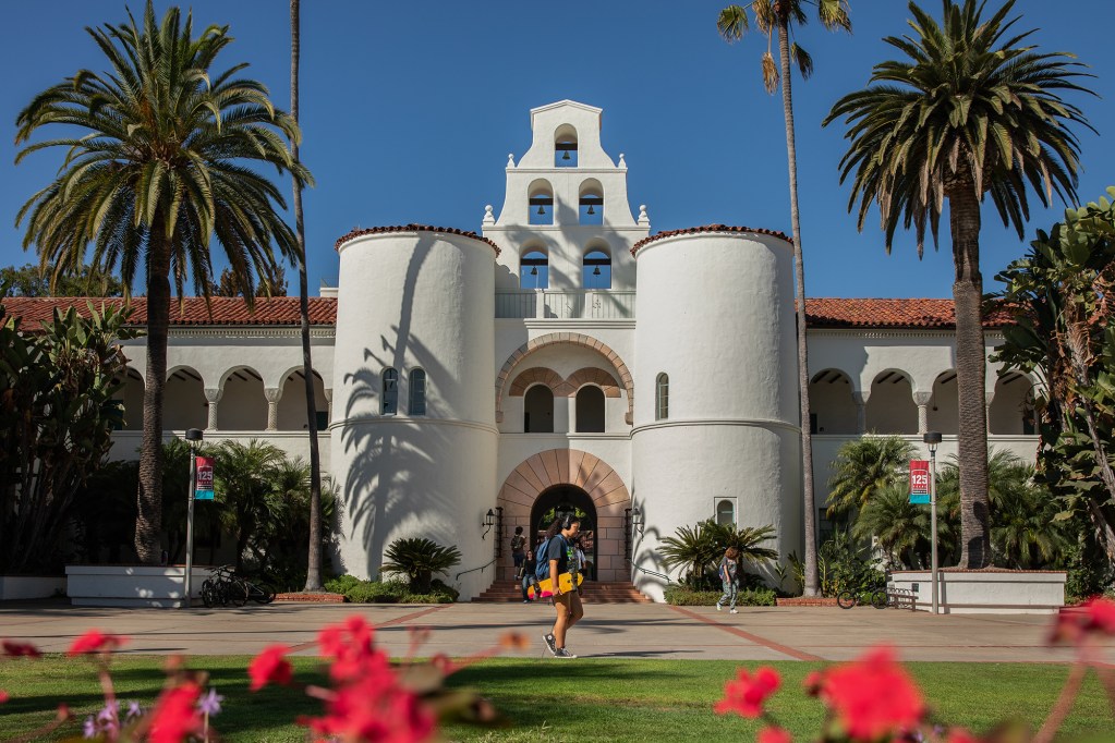 A person holding a skateboard walks by a white mission-style building surrounded by palm trees on a sunny and clear day.