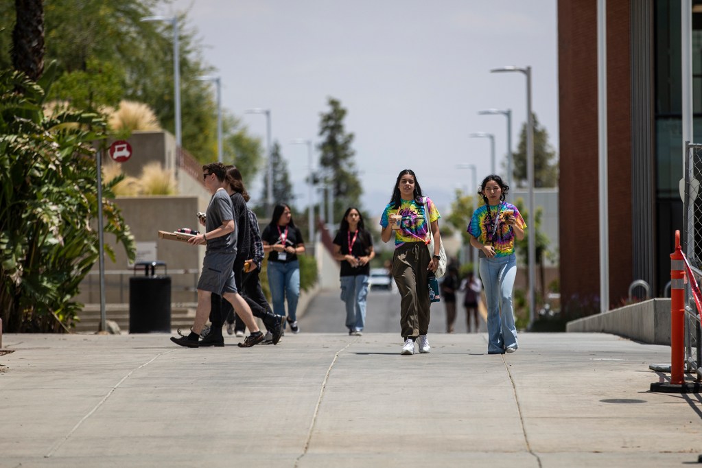 On a sunny day on a community college campus, students walk along a concrete path flanked by rows of light poles and between more buildings. Two students are wearing bright tie-dyed shirts and holding coffee cups as they walk.
