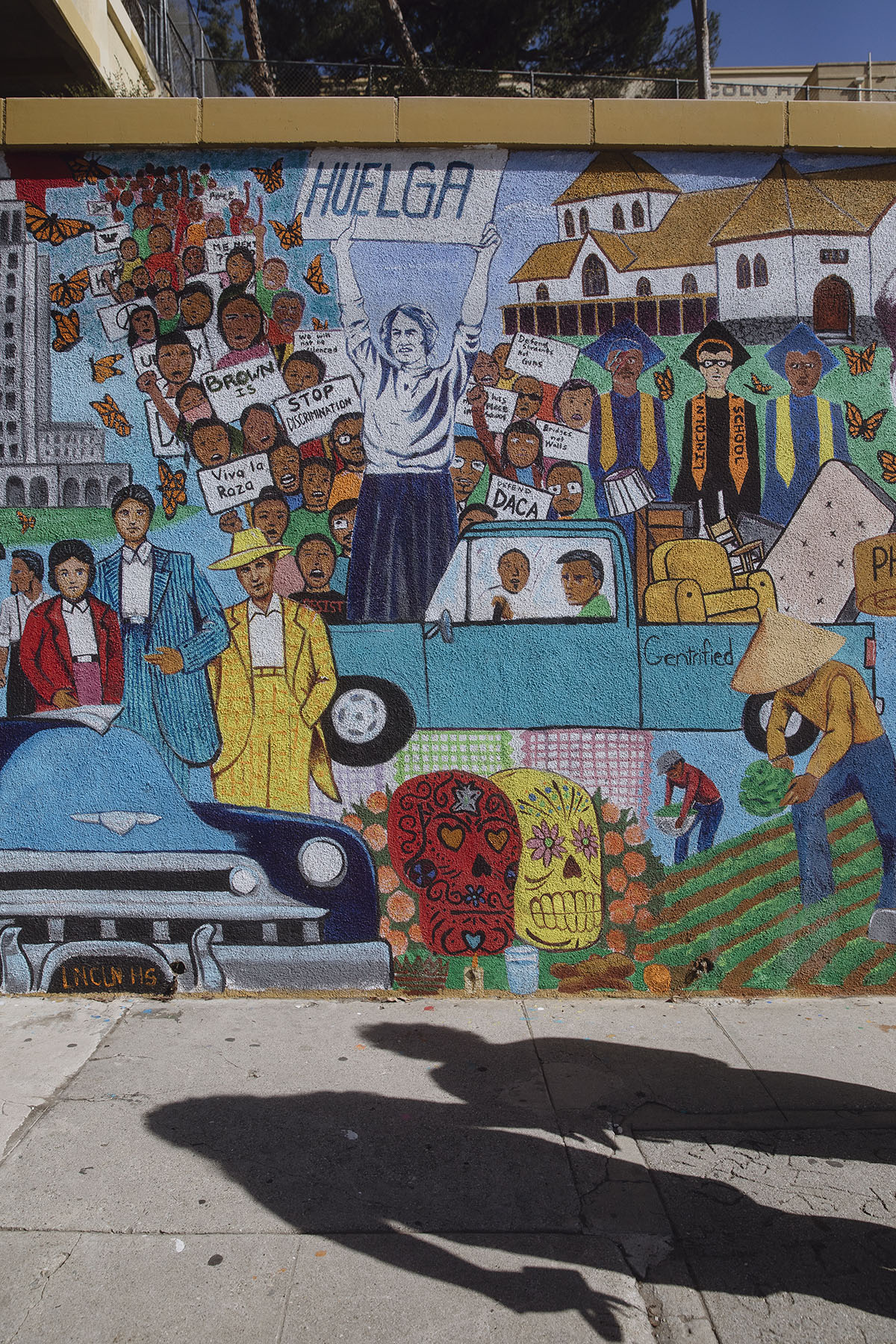 A colorful mural shows scenes from Chicano and immigrant activism, including raised fists, “HUELGA” signs, Day of the Dead skulls, and depictions of farmworkers and students.