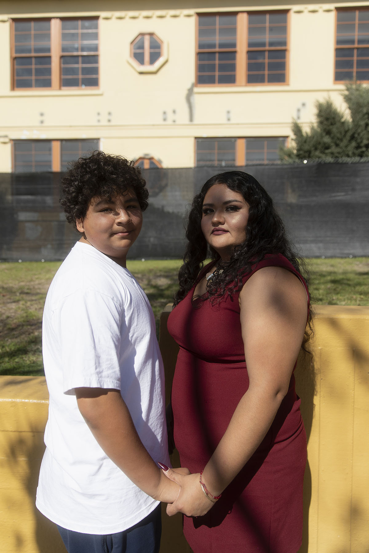Reyes and her teenage son Nathan stand side-by-side holding hands in front of a yellow school building, both looking directly at the camera.