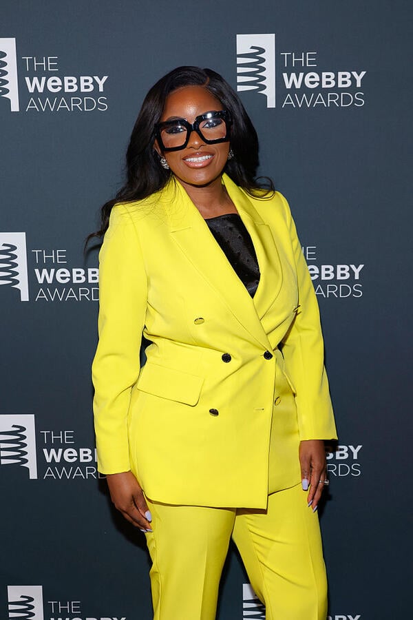 Jasmine Crocket, a Black woman with long black hair, is seen here wearing thick black glasses and a bright yellow-green suit.