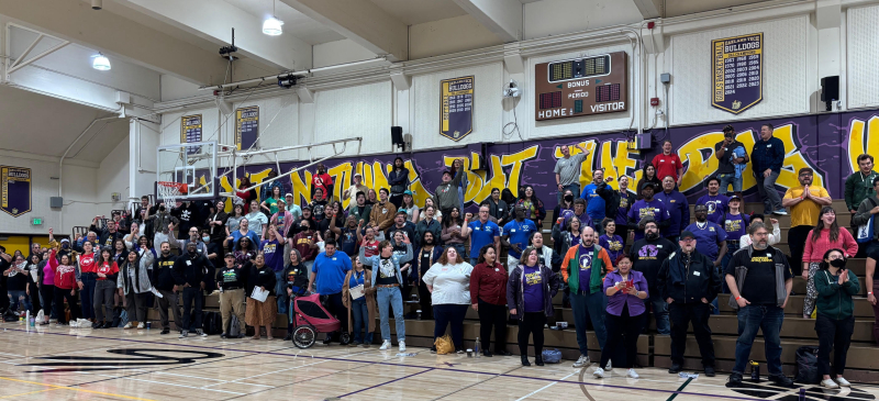 A large gathering of workers in purple, black, blue and other dark colored shirts. They're standing on the bleachers at a gymnasium.